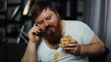 Kirli yağ adam görüşmeler hamburger yiyip kanepede oturan bira içmeyi Smartphone, sakallı. Yetersiz beslenme, gıda dağıtım, iletişim ve obezite kavramı