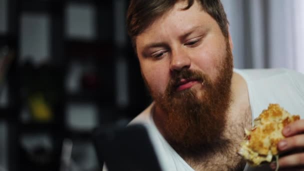 Толстяк с бородой печатает что-то в смартфоне, поедая бургер на диване. — стоковое видео