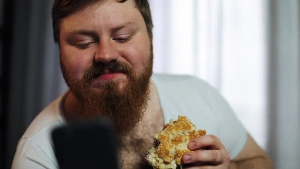Gordo barbudo escribe algo en su smartphone comiendo una hamburguesa en el sofá — Vídeo de stock