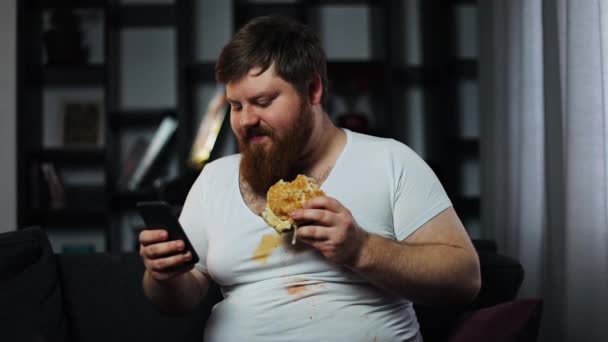 Fat bearded man types something in his smartphone eating a burger on the couch — Stock Video