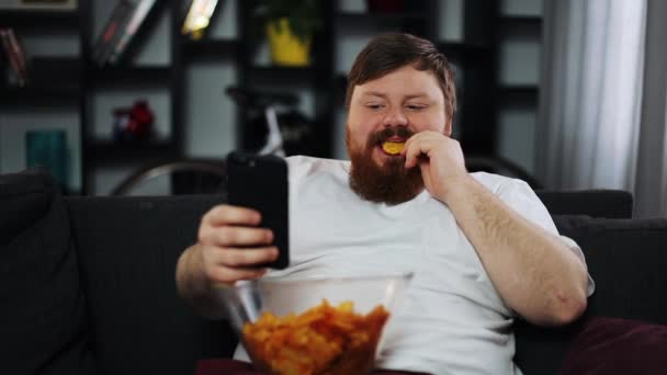 Dikke man glimlacht terwijl hij iets in zijn smartphone leest en potato chips eet — Stockvideo