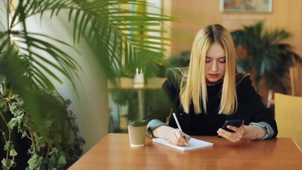 Young girl with smartphone doing homework sitting in cafe. Woman writes down something from her phone to the notebook — Stock Video