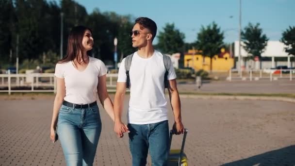 Feliz pareja joven va con el equipaje cerca del aeropuerto o la estación de tren. El concepto de viajes, vacaciones, vacaciones. Hora de verano, puesta de sol — Vídeo de stock