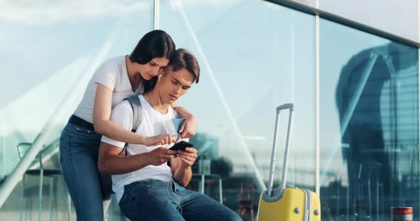 Szczęśliwa para w miłości za pomocą smartfonu. Młode małżeństwo zabawy w oczekiwaniu na lot na lotnisku. Kobieta jest dotknięcie ekranu tabletu — Wideo stockowe