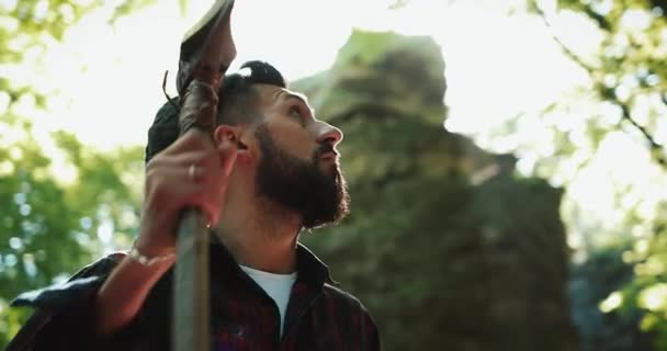 Lumberjack de pé com o machado na floresta, olhando em volta. Retrato de homem barbudo bonito com machado — Vídeo de Stock