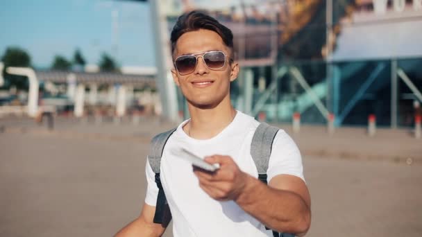 Um jovem e bonito homem de pé perto do aeroporto, olha para a câmera, sorri e mostra um trem de passagem de avião. O conceito de férias, descanso, viajar pelo mundo, trabalho e viagens — Vídeo de Stock