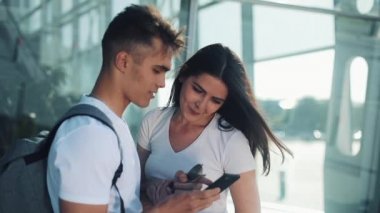 Yakışıklı güzel çift ayakta Havaalanı veya Tren İstasyonu yakınındaki. Onlar Smartphone onun parmak işaret. Genç çift mutlu ve memnun. Kayıtsızca giyinmiş.