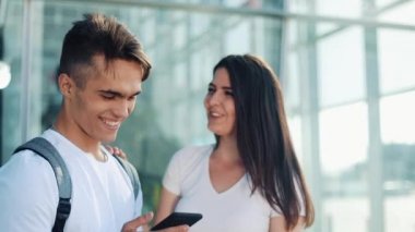 Yakışıklı güzel çift ayakta Havaalanı veya Tren İstasyonu yakınındaki. Onlar Smartphone onun parmak işaret. Genç çift mutlu ve memnun. Kayıtsızca giyinmiş.