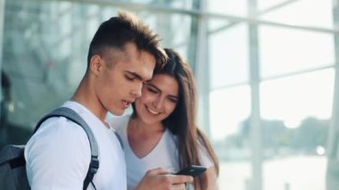 Yakışıklı güzel çift ayakta Havaalanı veya Tren İstasyonu yakınındaki. Onlar Smartphone onun parmak işaret. Genç çift mutlu ve memnun. Kayıtsızca giyinmiş.