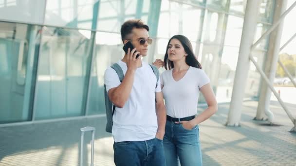 Attraktiva unga par stående nära flygplatsen. En man ringer en taxi eller ring av tjänsten. Kommunikation, teknik, smartphone koncept — Stockvideo