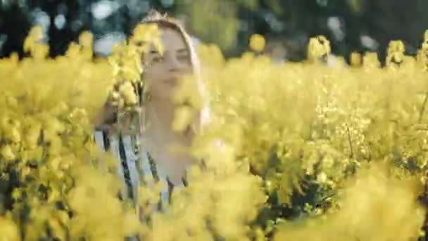 Chica bonita camina en el campo de violaciones, posando, y sonriendo a la cámara. Hermosa joven en el campo de la semilla de colza flores amarillas, día soleado — Vídeo de stock