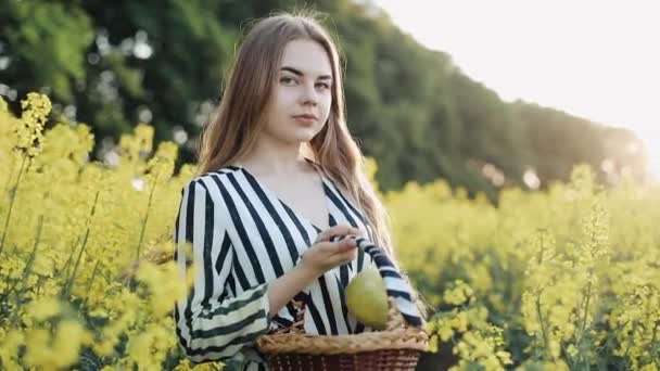 Menina bonita de pé no campo de sementes de colza flores amarelas. Ela segura um cesto de frutas em suas mãos. Uma jovem mulher em seu vestido segurar uma maçã verde — Vídeo de Stock