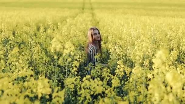Pretty young girl walks and turns around with waving hair in the rape field. Beautiful young woman in the field of rape seed yellow flowers, sunny day — Stock Video