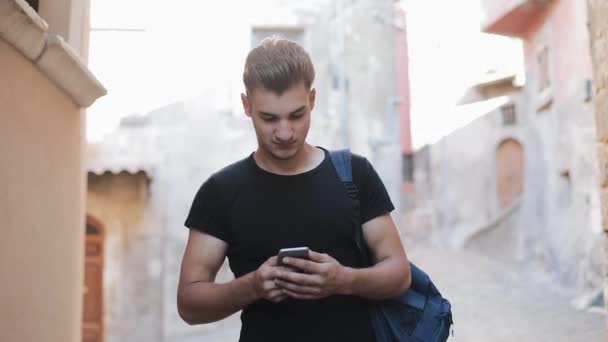 Junger Mann, der in der Altstadt spazieren geht und sein Smartphone benutzt. Männchen plaudert mit Freunden — Stockvideo