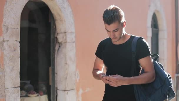 Un retrato de un joven guapo surfeando teléfono inteligente y caminando por la ciudad. Fondo soleado. Fondo borroso — Vídeo de stock