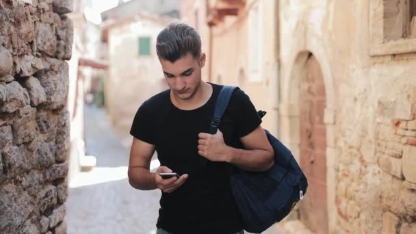 Atrakcyjne młody człowiek chodzą na starym mieście z plecakiem. Człowiek wybiera numer na telefonie komórkowym i czeka na odpowiedź — Wideo stockowe