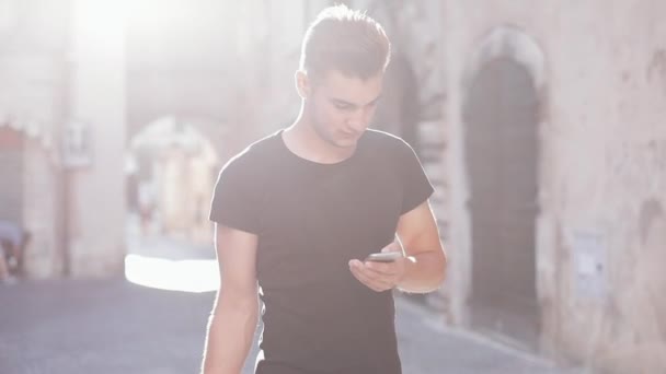Pemuda yang menarik berjalan di sekitar kota tua dengan ransel dan menggunakan ponsel. Sinar matahari — Stok Video