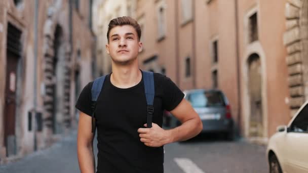 Молодой привлекательный турист с рюкзаком ходит по улице симпатичного города и оглядывается. На открытом воздухе — стоковое видео