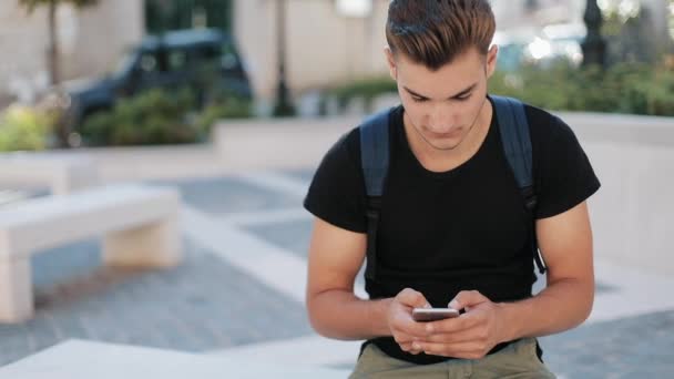 Atraktivní mladý muž s batohem na zádech telefonu sedí v staré město technologie zaneprázdněn portrét zblízka moderní pohledný smartphone ambiciózní mobilní slunce Zpomalený pohyb — Stock video