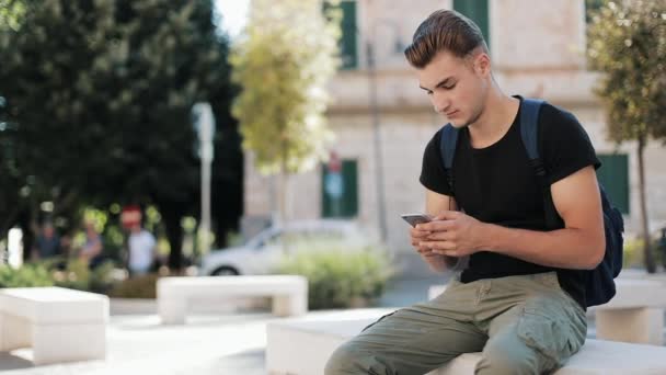 Atraktivní mladý muž s batohem na zádech telefonu sedí v staré město technologie zaneprázdněn portrét zblízka moderní pohledný smartphone ambiciózní mobilní slunce Zpomalený pohyb — Stock video