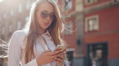 Beyaz gömlek giymiş ve şehir merkezi yaz internet iş teknoloji göz bahar mobil yavaş hareket sokak mutlu dışında yürürken smartphone kullanarak güzel genç iş kadını