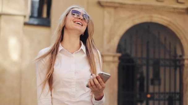 Bela jovem mulher de negócios vestindo camisa branca e usando smartphone enquanto caminha no centro da cidade negócios de internet de verão fora da tecnologia olho primavera móvel câmera lenta rua feliz — Vídeo de Stock