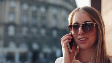 Güneşli şehir sokakları bina ve bir mobil cep telefonuyla konuşurken ofisinin duran genç iş kadını. Cep telefonu kullanarak güneş gözlüğü ile çekici mutlu kız açık havada, gün batımı