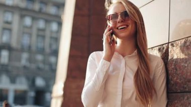 Güneşli şehir sokakları bina ve bir mobil cep telefonuyla konuşurken ofisinin duran genç iş kadını. Cep telefonu kullanarak güneş gözlüğü ile çekici mutlu kız açık havada, gün batımı