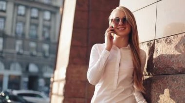 Güneşli şehir sokakları bina ve bir mobil cep telefonuyla konuşurken ofisinin duran genç iş kadını. Cep telefonu kullanarak güneş gözlüğü ile çekici mutlu kız açık havada, gün batımı