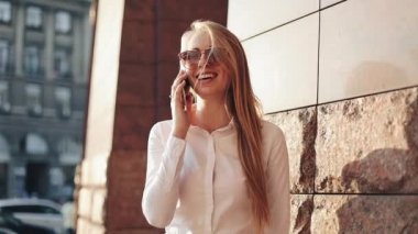 Güneşli şehir sokakları bina ve bir mobil cep telefonuyla konuşurken ofisinin duran genç iş kadını. Cep telefonu kullanarak güneş gözlüğü ile çekici mutlu kız açık havada, gün batımı