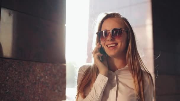 Eine junge Geschäftsfrau steht in der Nähe eines Bürogebäudes an den sonnigen Straßen der Stadt und telefoniert mit einem Mobiltelefon. attraktive glückliche Mädchen mit Sonnenbrille mit Handy im Freien, Sonnenuntergang — Stockvideo