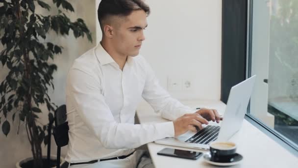 Χαρούμενος νεαρός επιχειρηματίας που εργάζεται σε ένα φορητό υπολογιστή στην καφετέρια κοντά στο παράθυρο. Εκείνος τελειώνει το έργο του και κλείνει το laptop. Επαγγελματικά, καριέρας, επιτυχία, freelancer έννοια αναζήτηση θέσεων εργασίας — Αρχείο Βίντεο