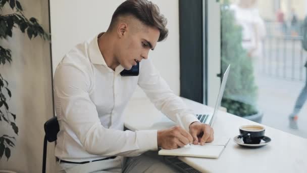 Средний снимок концентрированного молодого бизнесмена, делающего заметки в блокноте во время разговора по телефону во время кофе-брейка — стоковое видео