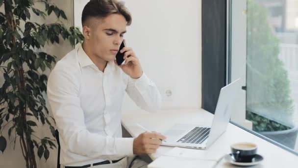 Middellange schot van geconcentreerde jonge zakenman die aantekeningen in notitieblok wanneer op telefoon te praten tijdens de koffiepauze — Stockvideo