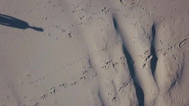Havadan çekim. Üç erkek çölde birleşirler. Güneşin gölgeleri kum atar. Üstten görünüm