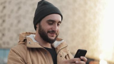 Shopping mall yakınındaki sokakta Smartphone kullanan mutlu genç sakallı adam. O bir sonbahar ceket ve örme şapka giyiyor. İletişim, online alışveriş, sohbet, sosyal ağ kavramı