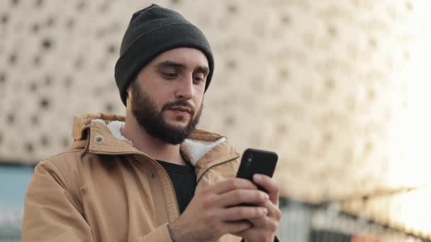 Homem barba jovem feliz usando smartphone na rua perto de shopping center. Ele está usando uma jaqueta de outono e chapéu de malha. Comunicação, compras online, bate-papo, conceito de redes sociais — Vídeo de Stock