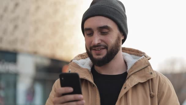 Hombre barba joven feliz usando teléfono inteligente en la calle cerca del centro comercial. Lleva una chaqueta de otoño y un sombrero de punto. Comunicación, compras en línea, chat, concepto de redes sociales — Vídeos de Stock