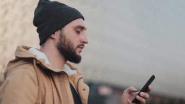 Shopping mall yakınındaki sokakta yürürken smartphone kullanan mutlu genç sakallı adam. O bir sonbahar ceket ve örme şapka giyiyor. İletişim, online alışveriş, sohbet, sosyal ağ kavramı