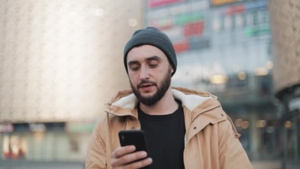 Gelukkig jonge baard man met smartphone wandelen in de straat in de buurt van shopping mall. Hij draagt een herfst jas en gebreide muts. Communicatie, online winkelen, chat, sociale netwerken concept — Stockvideo