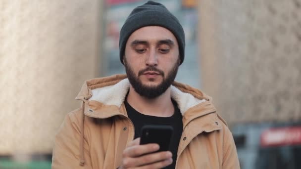 Щасливий молодий бородатий чоловік використовує смартфон, що йде вулицею біля торгового центру. Він одягнений в осінню куртку і в'язаний капелюх. Зв'язок, онлайн-покупки, чат, концепція соціальних мереж — стокове відео