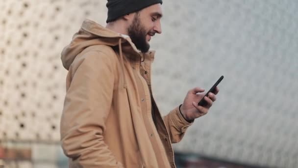 Muž šťastný mladý vousy pomocí smartphone chůzi na ulici nedaleko nákupního centra. Má na sobě podzimní bunda a čepice pletená. Komunikace, nakupování on-line, chat, sociální sítě koncepce — Stock video