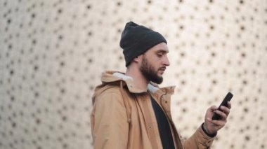 Shopping mall yakınındaki sokakta yürürken smartphone kullanan mutlu genç sakallı adam. O bir sonbahar ceket ve örme şapka giyiyor. İletişim, online alışveriş, sohbet, sosyal ağ kavramı