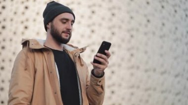 Shopping mall yakınındaki sokakta Smartphone kullanan mutlu genç sakallı adam. O bir sonbahar ceket ve örme şapka giyiyor. İletişim, online alışveriş, sohbet, sosyal ağ kavramı