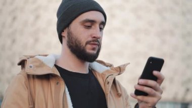 Shopping mall yakınındaki sokakta Smartphone kullanan mutlu genç sakallı adam. O bir sonbahar ceket ve örme şapka giyiyor. İletişim, online alışveriş, sohbet, sosyal ağ kavramı