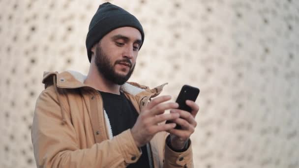 Człowiek szczęśliwy młody brodę przy użyciu smartfona na ulicy w pobliżu centrum handlowego. Ma na sobie jesień marynarka i czapka. Komunikacji, zakupy online, czat, social networking koncepcja — Wideo stockowe