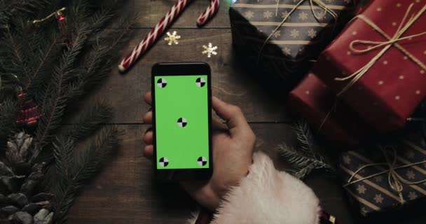 Rukou Santa Claus drží černý smartphone s zeleným plátnem. Jiné ruce umístění některé současné krabic na stole. Chromatický klíč. Pohled shora. Vánoce Nový rok internetové objednávky koncept — Stock video