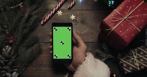 녹색 스크린 블랙 스마트폰 들고 산타 클로스의 손. 다른 손은 테이블에 몇 가지 있는 상자를 배치. 크로마 키입니다. 최고의 볼 수 있습니다. 크리스마스 새 해 인터넷 주문 개념 — 비디오