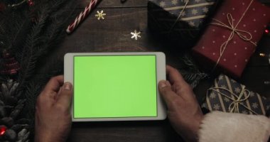 Claus Santa yeşil ekran beyaz tablet bilgisayarınızla tutan eller. Bazı mevcut kutuları masaya yerleştirerek diğer eller. Chroma anahtar. Üstten görünüm. Yılbaşı yeni yıl Internet sipariş kavramı
