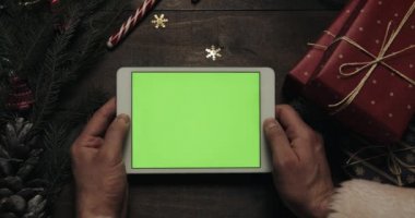 Claus Santa yeşil ekran beyaz tablet bilgisayarınızla tutan eller. Bazı mevcut kutuları masaya yerleştirerek diğer eller. Chroma anahtar. Üstten görünüm. Yılbaşı yeni yıl Internet sipariş kavramı